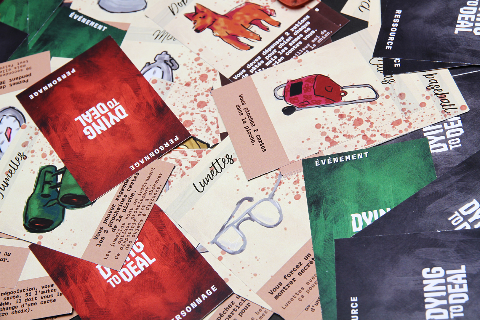 photographie de cartes éparpillées, on peut lire dying to deal au dos de certaines, sur d'autres, on voit une illustration, par exemple un chien, un sac cabas, des lunettes...