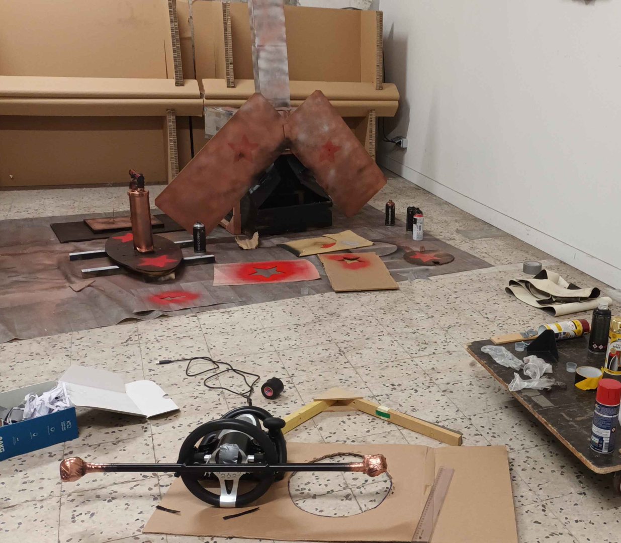 Photographie d'une structure de carton en forme de chaudière à charbon, d'une pompe, d'un volant, décorés en noir, rouge et marron, autour d'outils de peinture