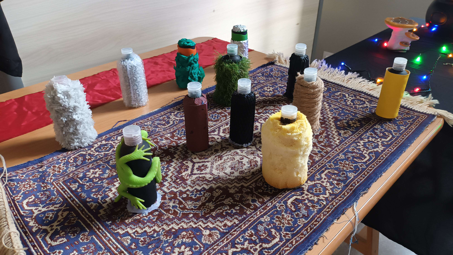 Photographie d'une dizaine de bouteilles de différentes matières (mousse, laine, papier crépon, cordelette, papier de verre, etc), un lézard en caoutchouc est enroulé autour de l'une d'elles